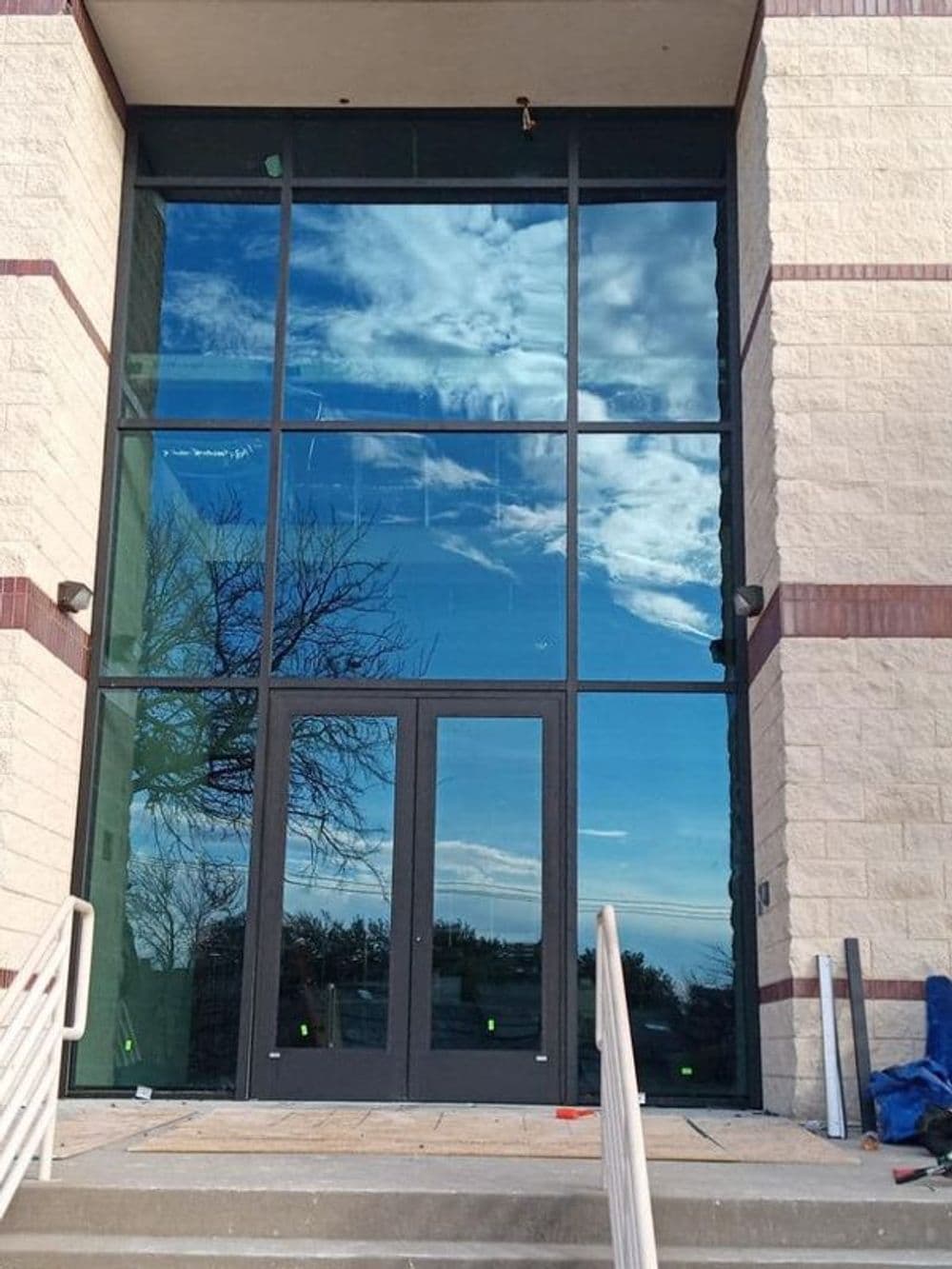 Modern glass entrance with reflections of clouds and trees, featuring two black doors.